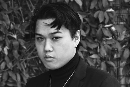 A serious looking young man in a turtleneck sweater and jacket wears a necklace and poses for a black and white photo in front of foliage.